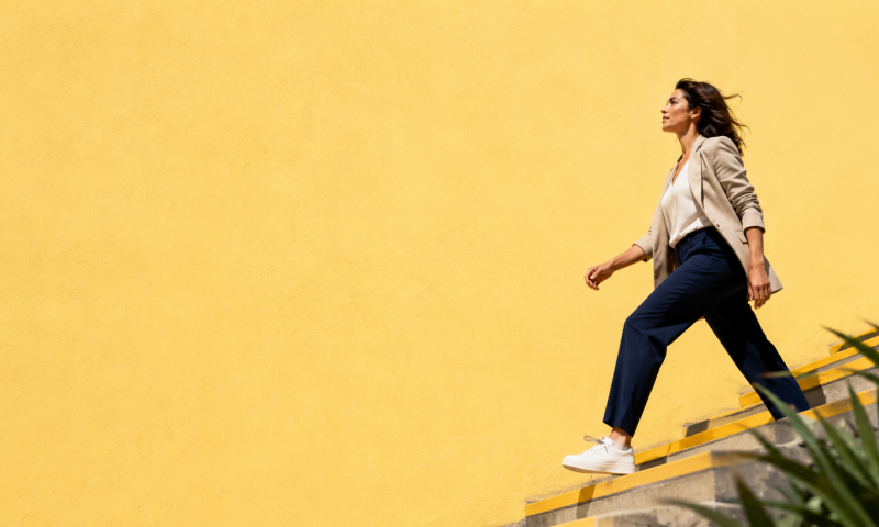 Editorial image of a professional woman walking upward on bright yellow steps against a vibrant yellow wall, representing the shift from useful production to confident ownership.