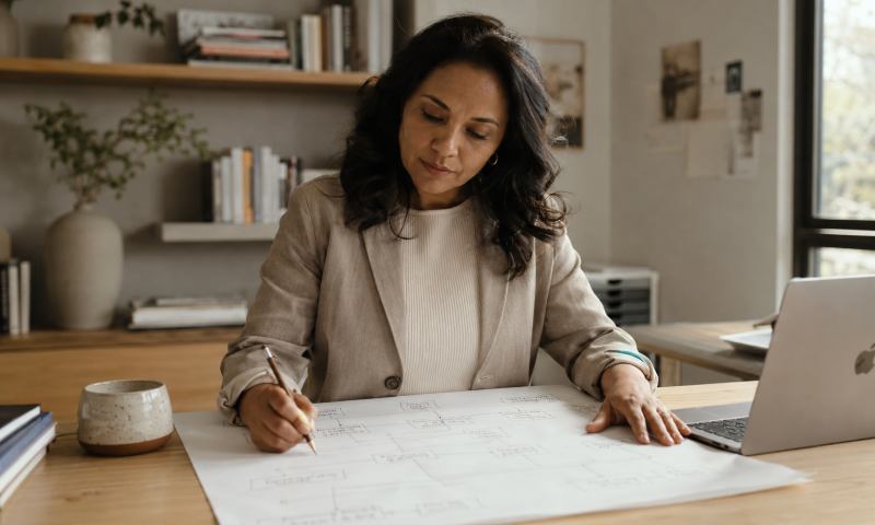 Woman studying a hand-drawn flowchart at a light wood desk in a warm modern office, with a pencil, coffee cup and MacBook nearby.